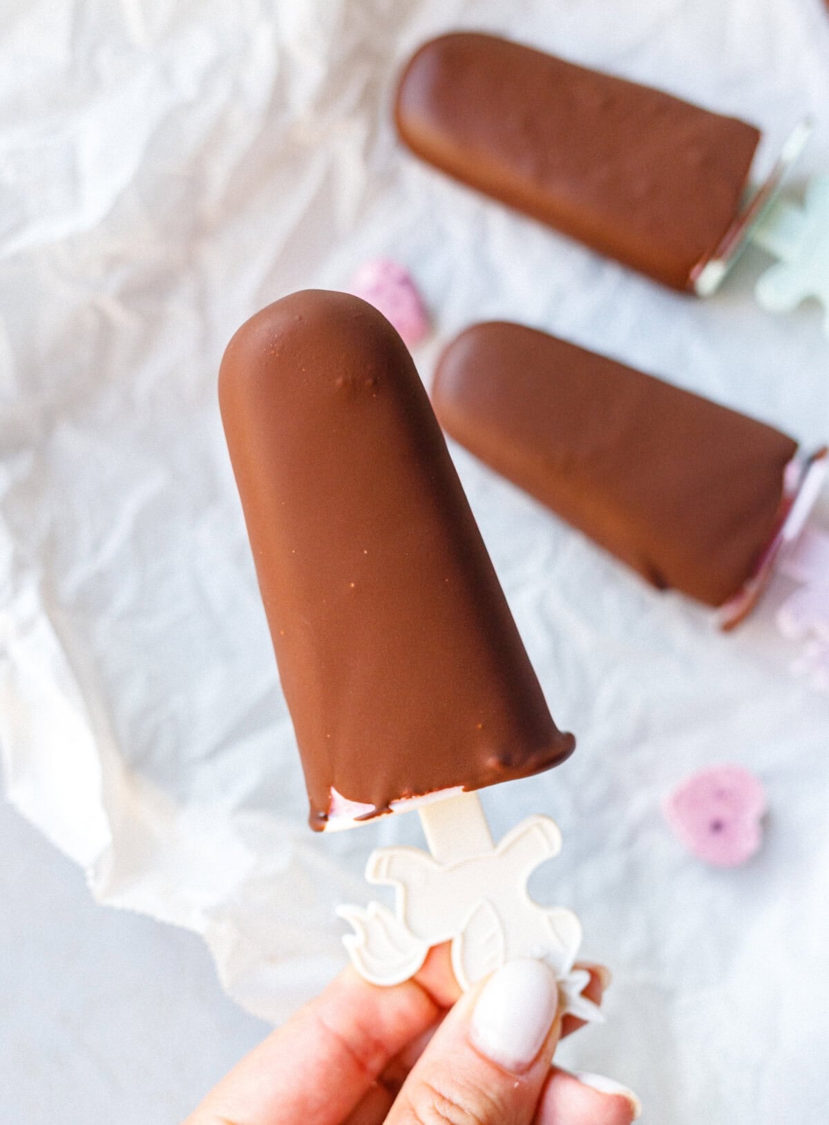 Hand holding a chocolate-covered cottage cheese popsicle on a unicorn stick over parchment paper