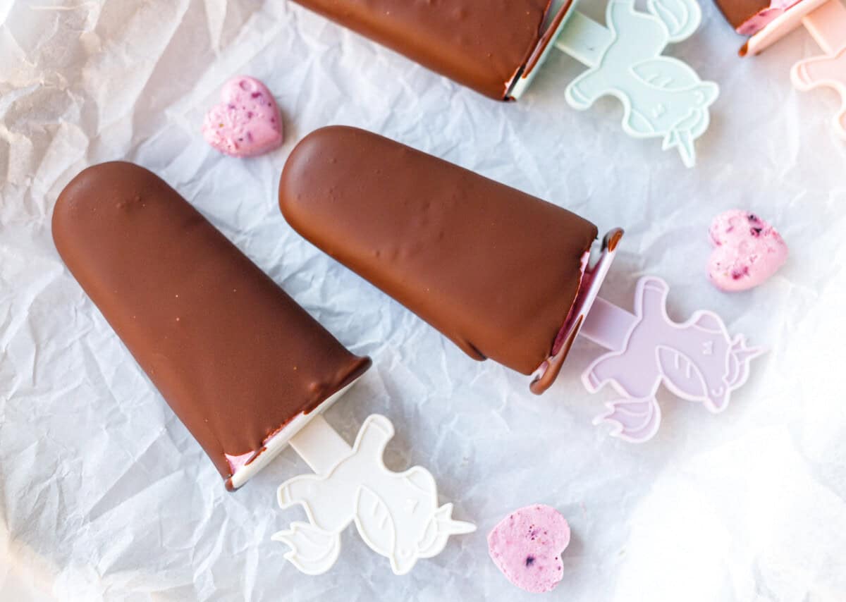 Two chocolate-dipped cottage cheese popsicles on parchment with pink berry hearts and pastel unicorn sticks