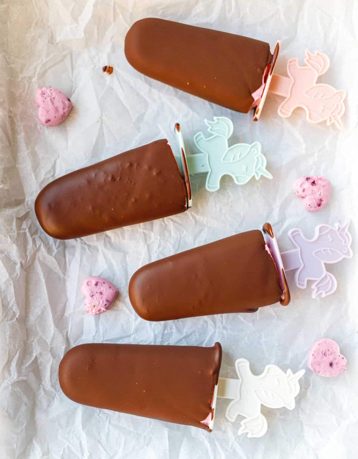 Four chocolate-coated cottage cheese popsicles with colorful unicorn sticks on crinkled parchment