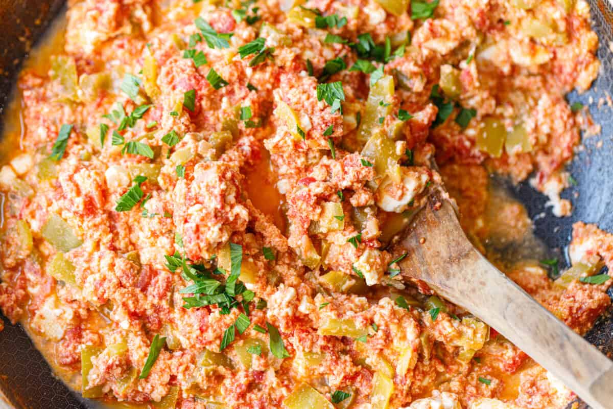 Skillet of peppers, tomatoes and eggs with feta and parsley, wooden spoon resting in the pan
