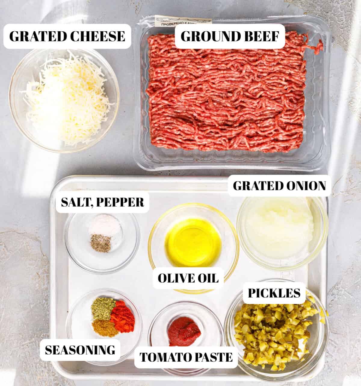 Ingredients for ground beef tortilla pinwheels — grated cheese, ground beef, grated onion, chopped pickles, tomato paste, olive oil, salt, pepper and spices in glass bowls on a metal tray.
