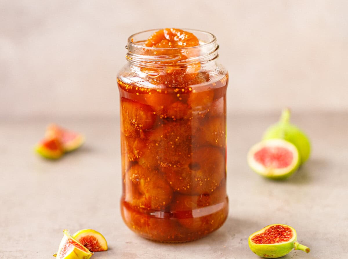 Glass jar filled with whole fig preserves in amber syrup, with fresh green figs cut in half on the table