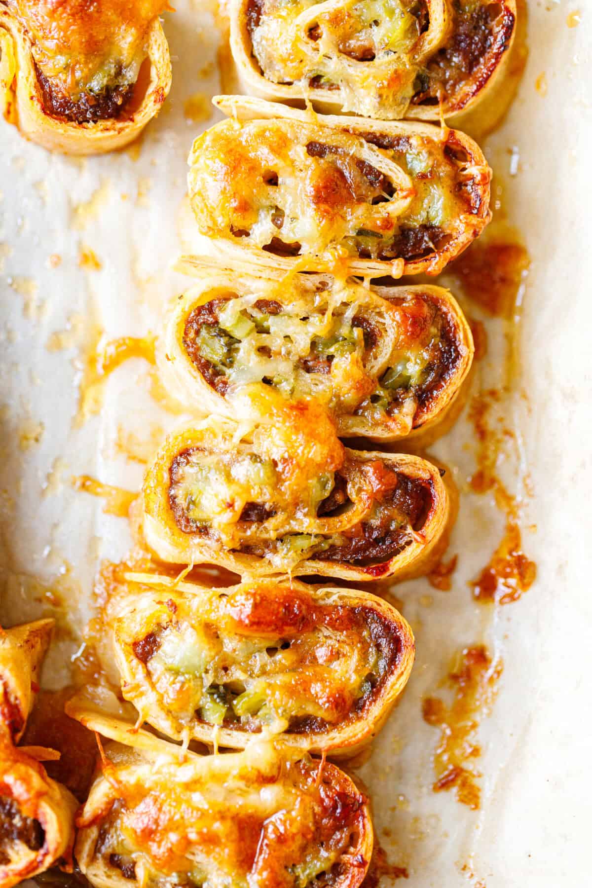 Baked ground beef tortilla pinwheels in a row on parchment, cheese pulling between them, showing the spiral of beef and chopped pickles inside.
