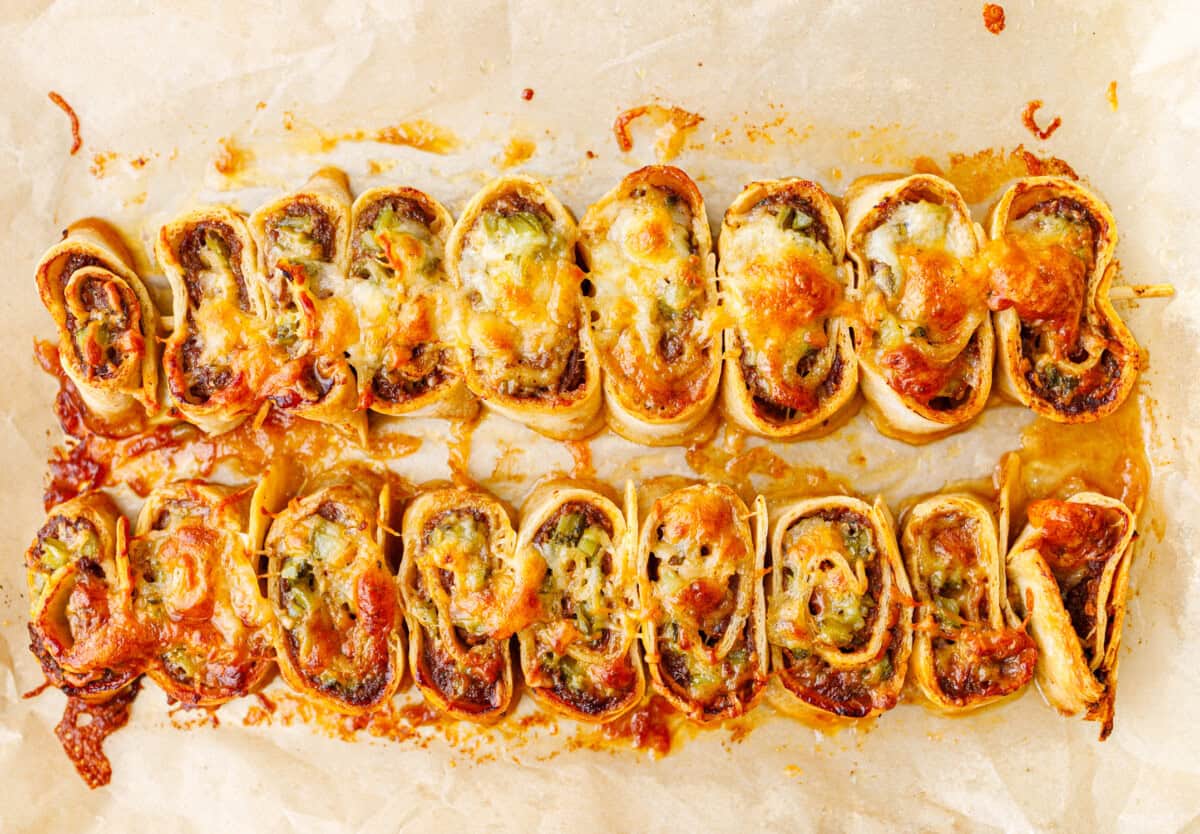 Two rows of baked ground beef tortilla pinwheels on parchment paper, tops browned and bubbling with melted cheese.