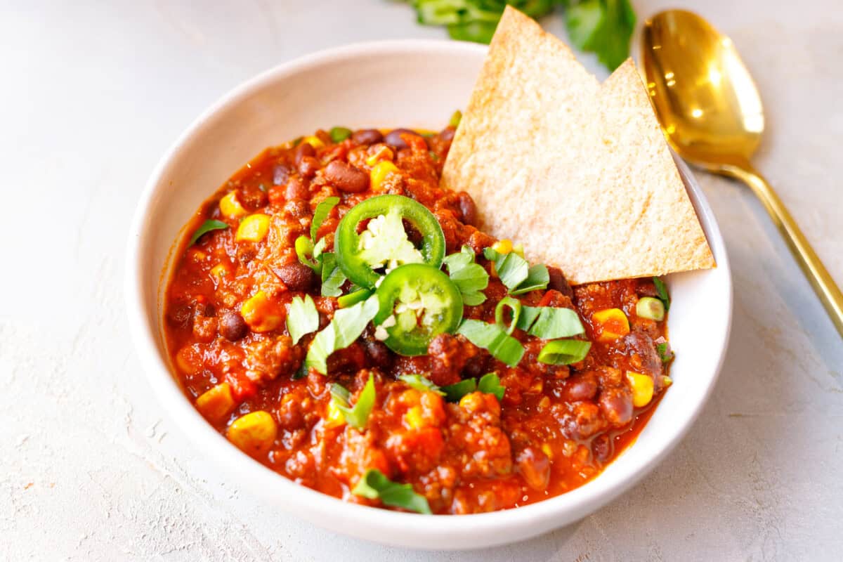 A bowl of easy crockpot chili with beef, beans and corn, topped with sliced jalapeño, herbs and a tortilla chip, with a gold spoon beside it.