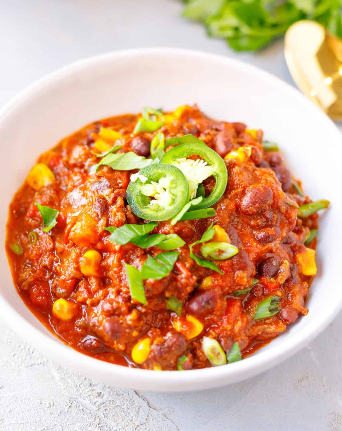 A white bowl of slow cooker chili with ground beef, kidney and black beans and corn, topped with jalapeño slices, green onion and parsley.