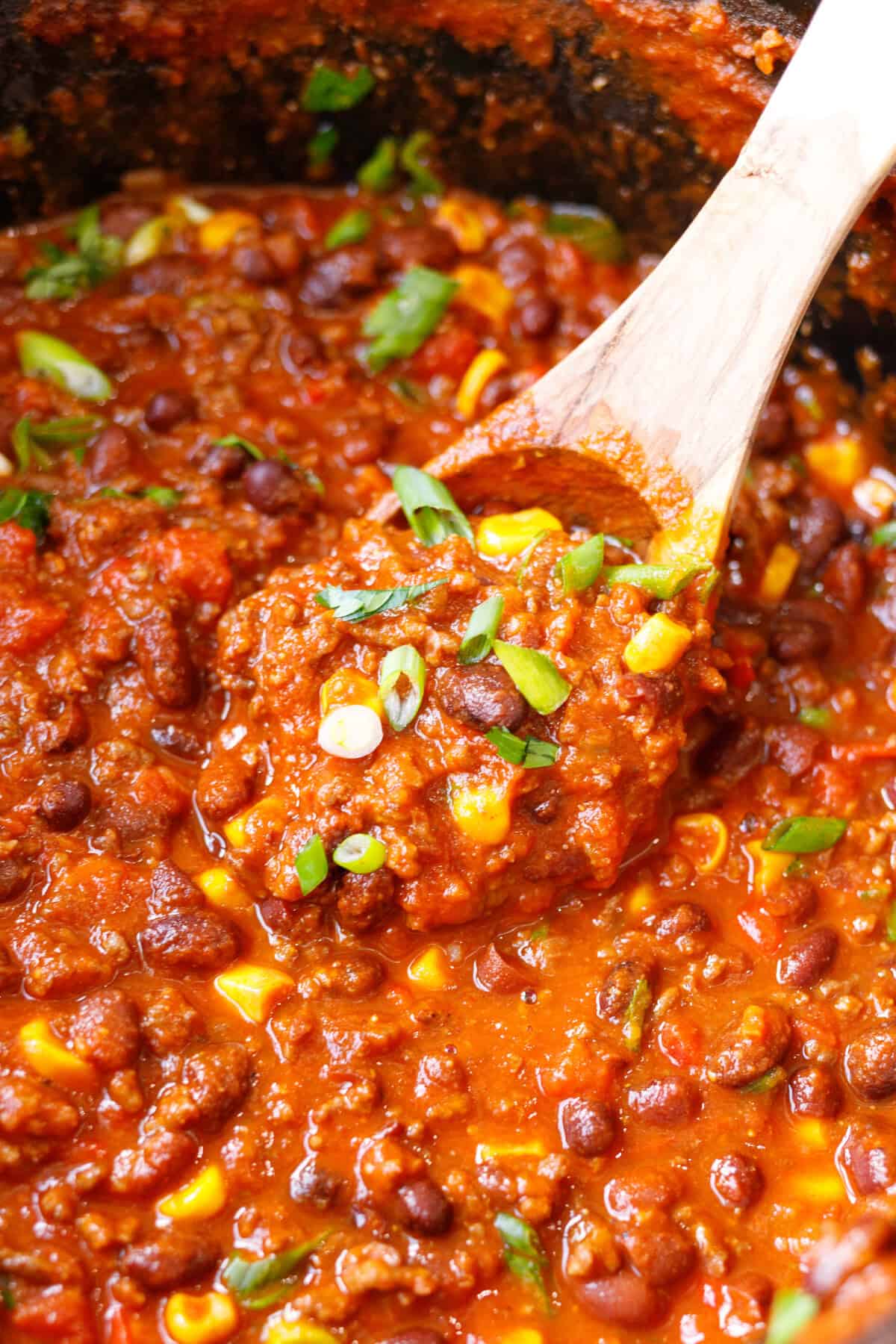 A wooden spoon lifting crockpot chili with ground beef, kidney and black beans, corn and green onion.