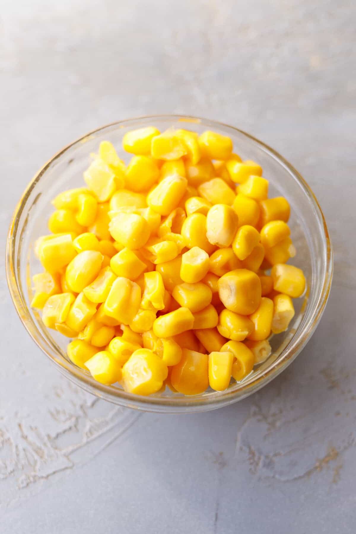 A small glass bowl of drained canned sweet corn on a gray surface.