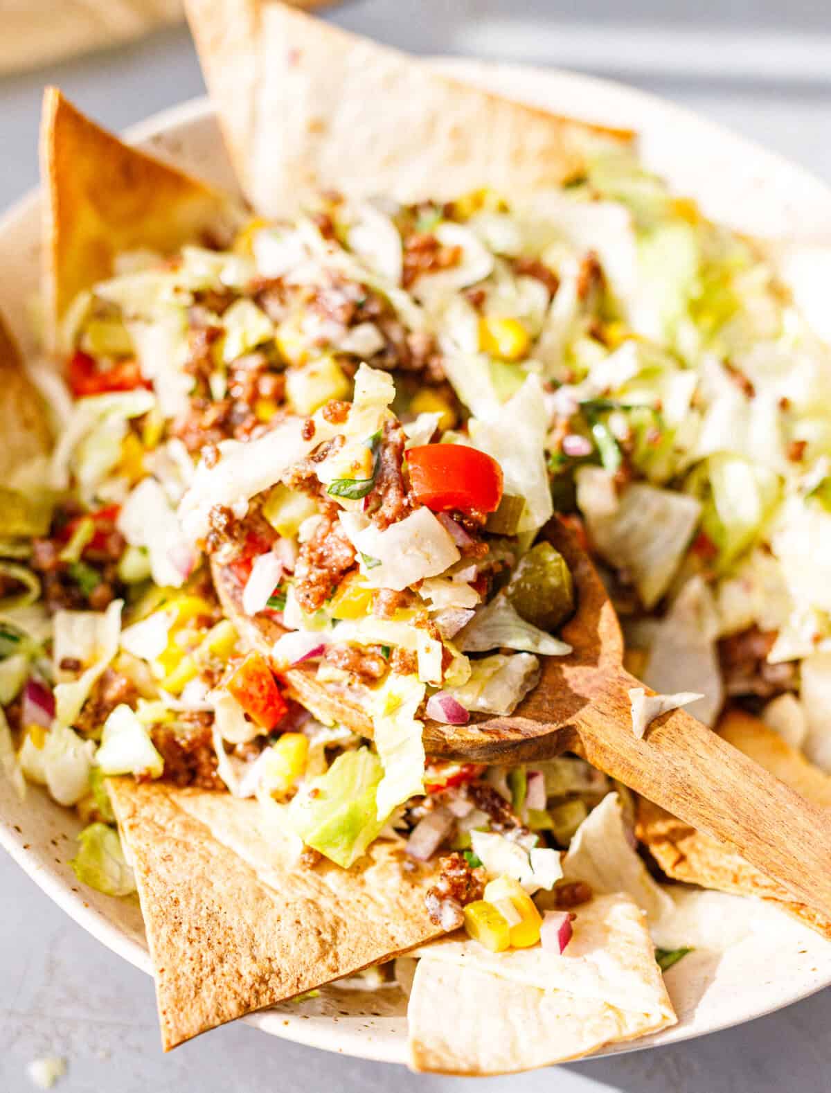 A wooden spoon lifting tossed ground beef taco salad with iceberg, tomato, pickles and corn from a bowl lined with tortilla chips.