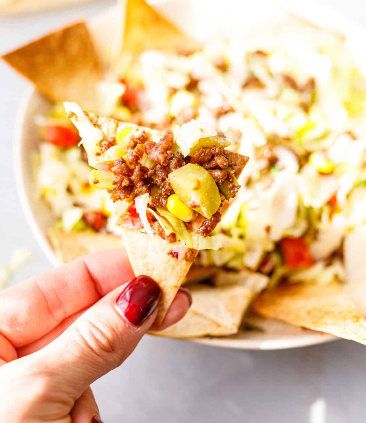 A hand holding a homemade tortilla chip topped with ground beef taco salad, with the salad bowl behind it.