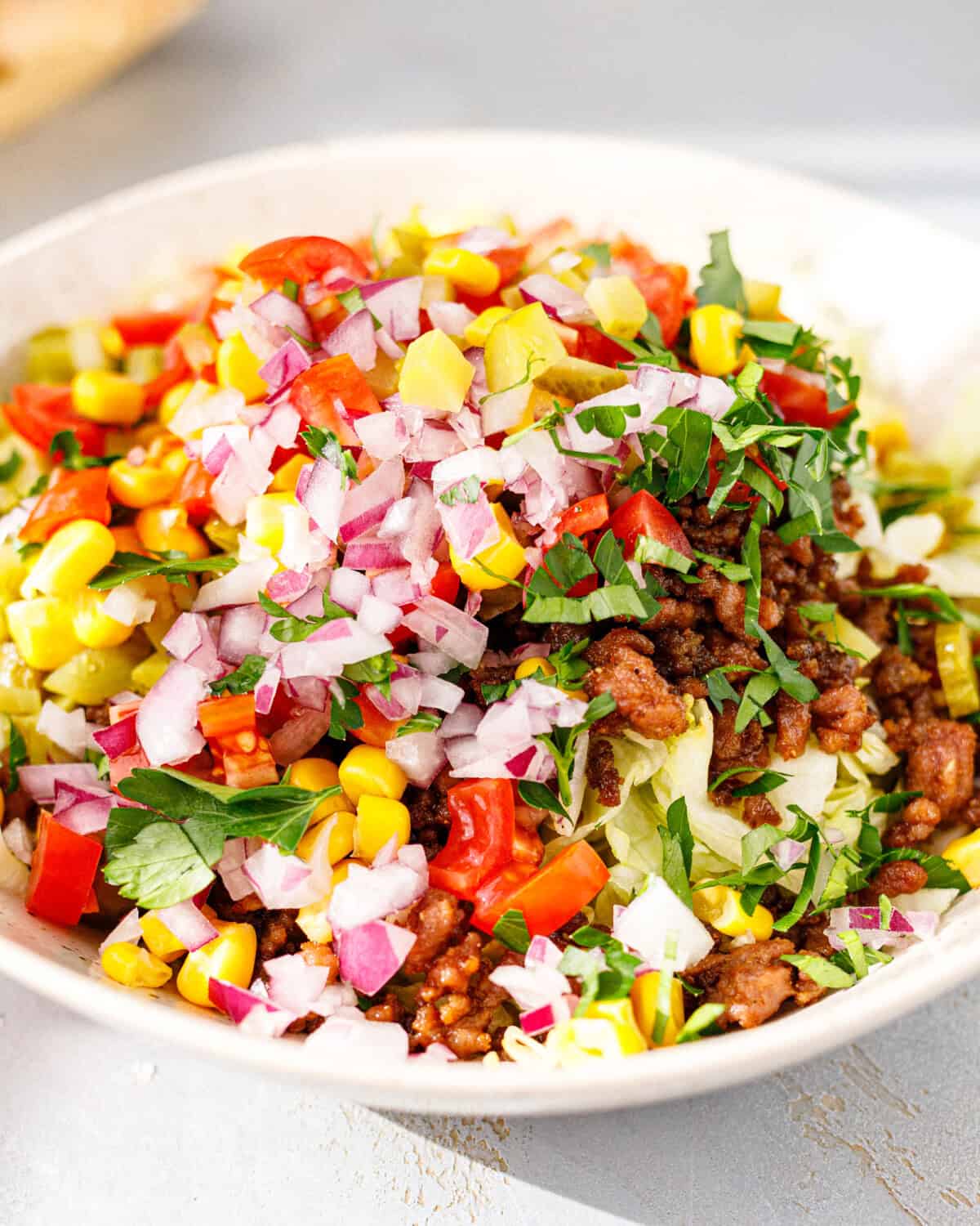 A side view of a taco salad bowl with seasoned ground beef, iceberg, corn, cherry tomatoes, red onion, pickles and fresh herbs.