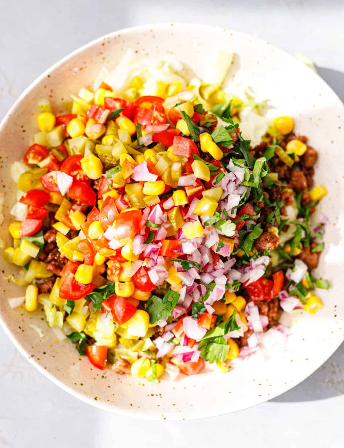 A colorful taco salad bowl with ground beef, iceberg, sweet corn, cherry tomatoes, pickles, red onion and fresh cilantro.