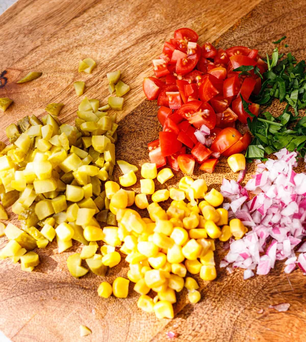 Chopped pickles, sweet corn, cherry tomatoes, red onion and cilantro on a wooden cutting board for taco salad.