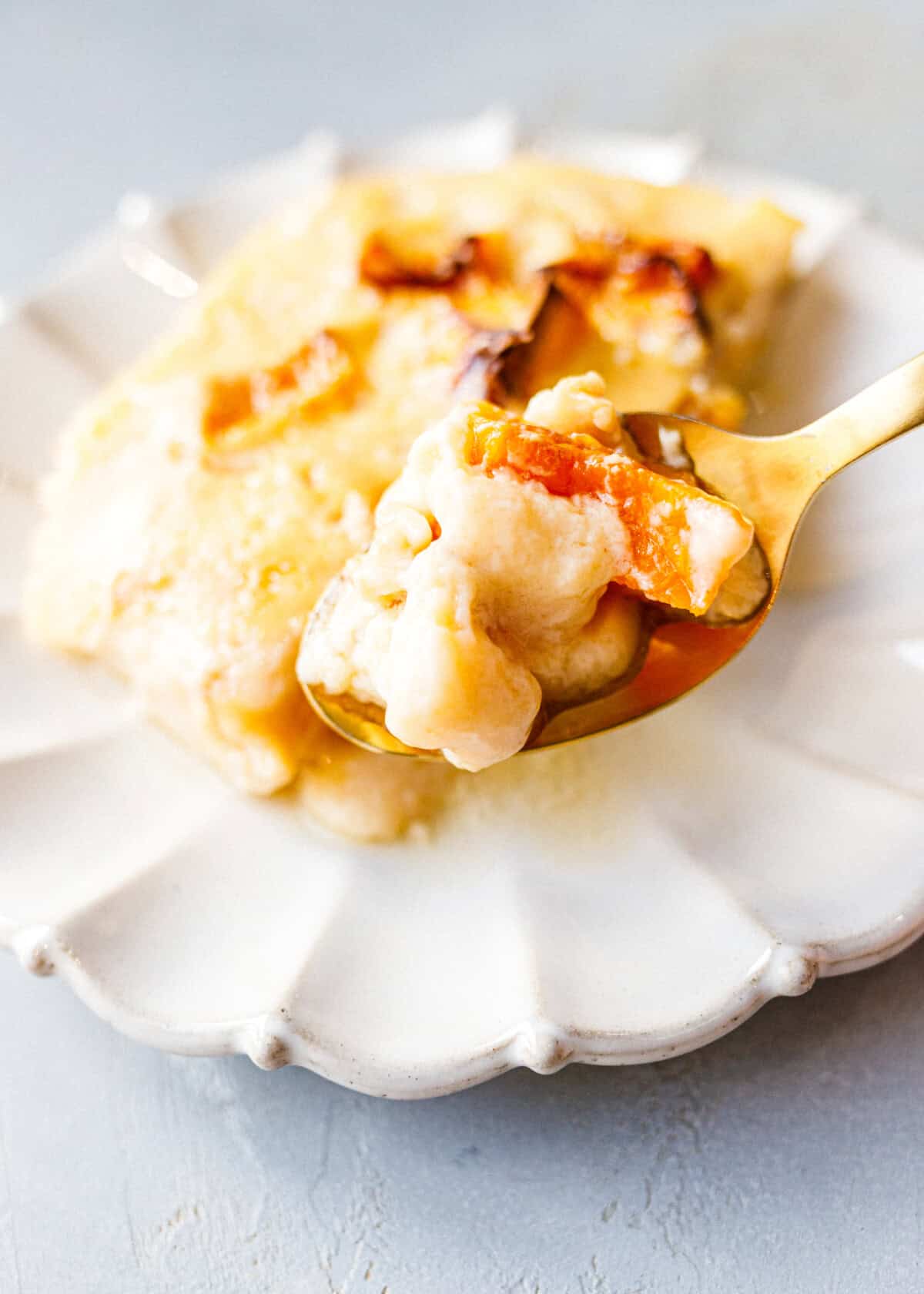 A gold spoon holding a bite of baked pumpkin custard with soft roasted pumpkin pieces, in front of a slice on a scalloped white plate.