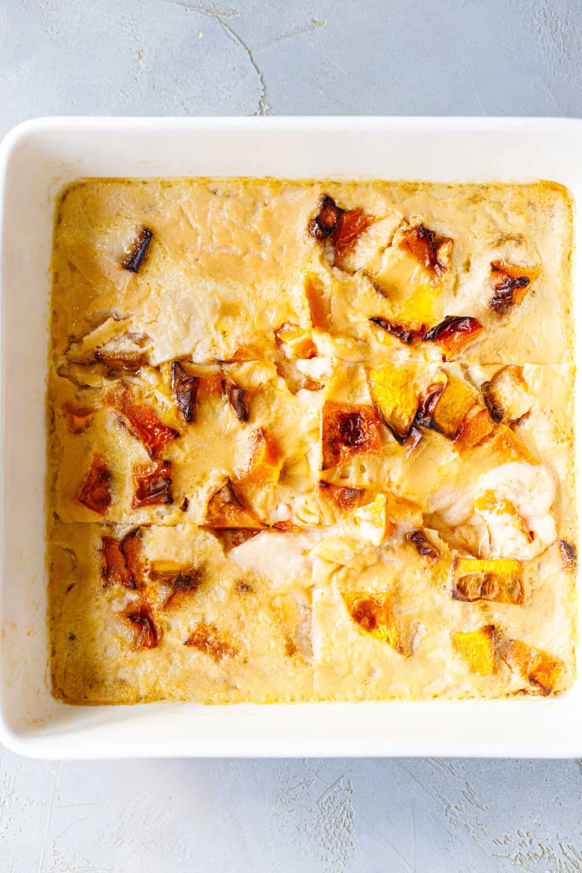 Overhead view of baked pumpkin custard in a square white baking dish, cut into squares, with golden roasted pumpkin pieces.