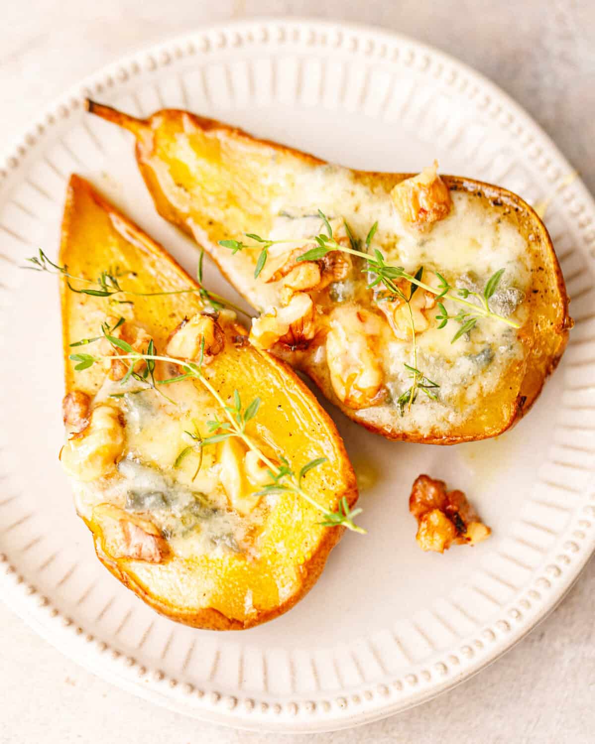 Two savory baked pear halves with melted blue cheese, walnuts and fresh thyme on a white ceramic plate