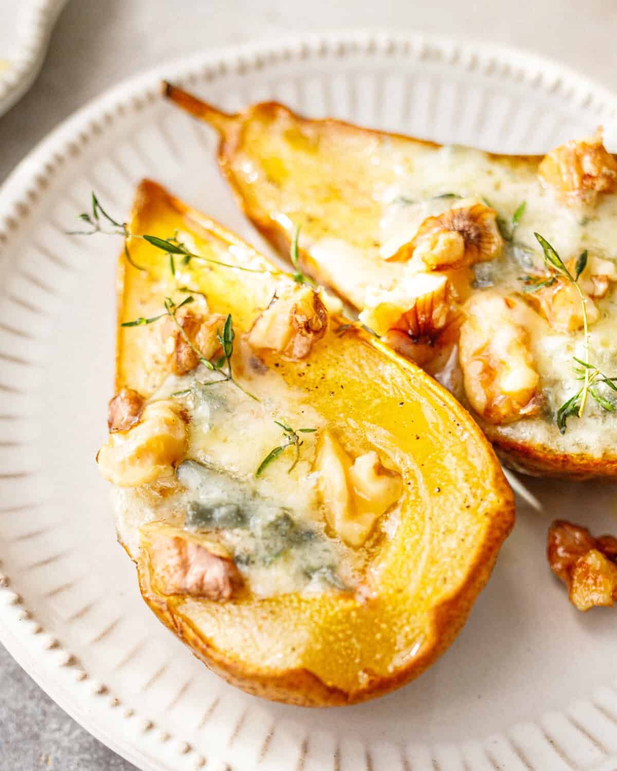 Close-up of a baked pear half with melted blue cheese, toasted walnuts and fresh thyme on a white plate