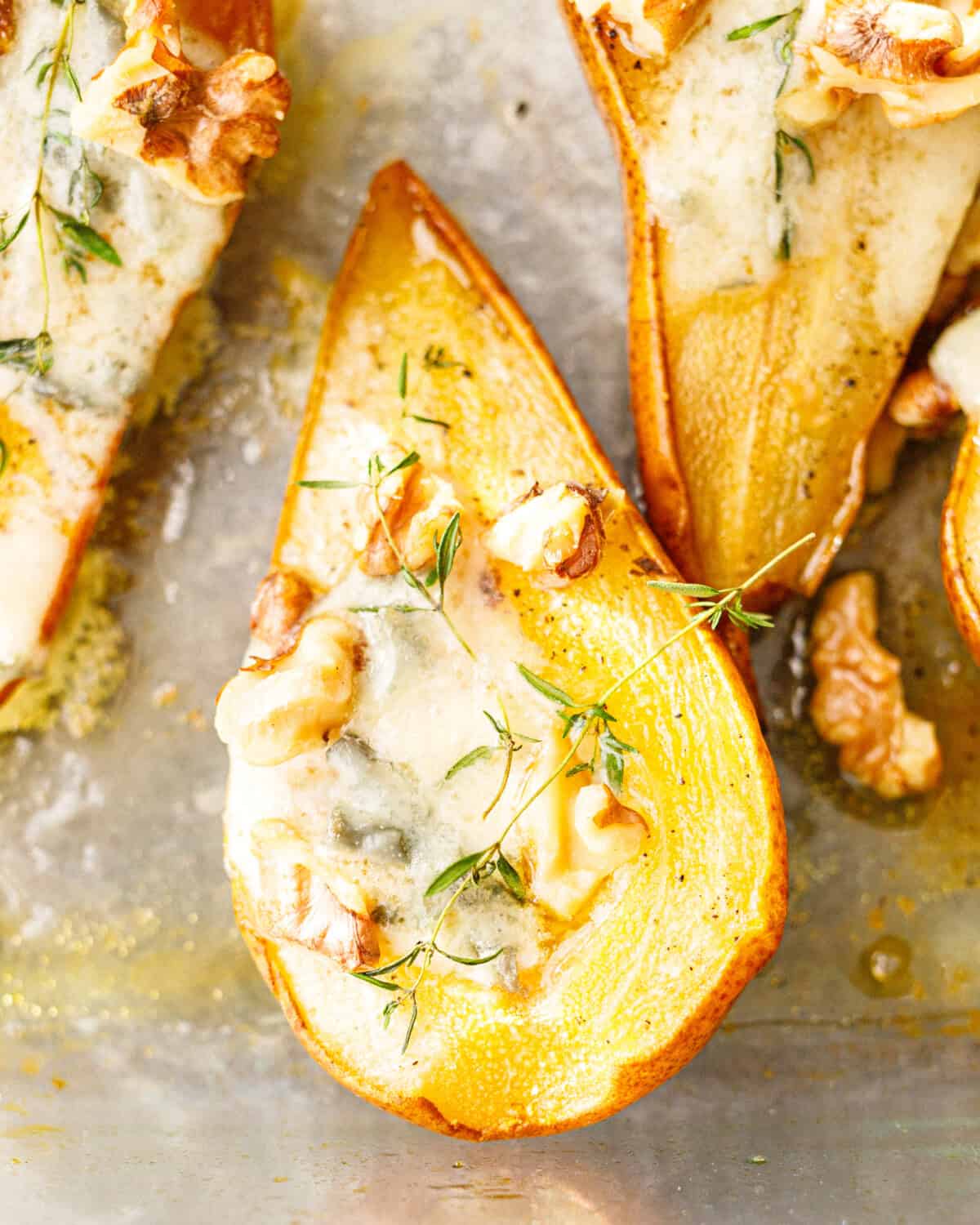 Close-up of a baked pear half filled with melted blue cheese, toasted walnuts and thyme in a baking dish