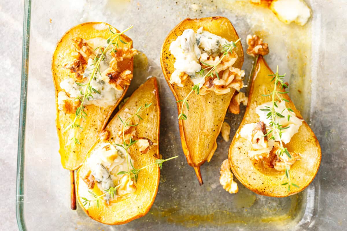 Overhead view of four baked pear halves with crumbled blue cheese, toasted walnuts and fresh thyme in a glass baking dish