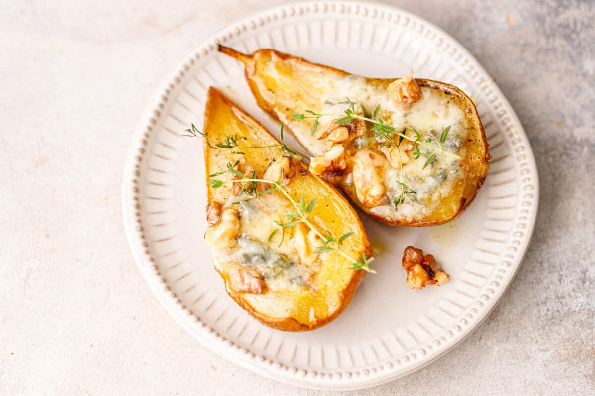 Two roasted pear halves with melted blue cheese, walnuts and fresh thyme on a white ceramic plate
