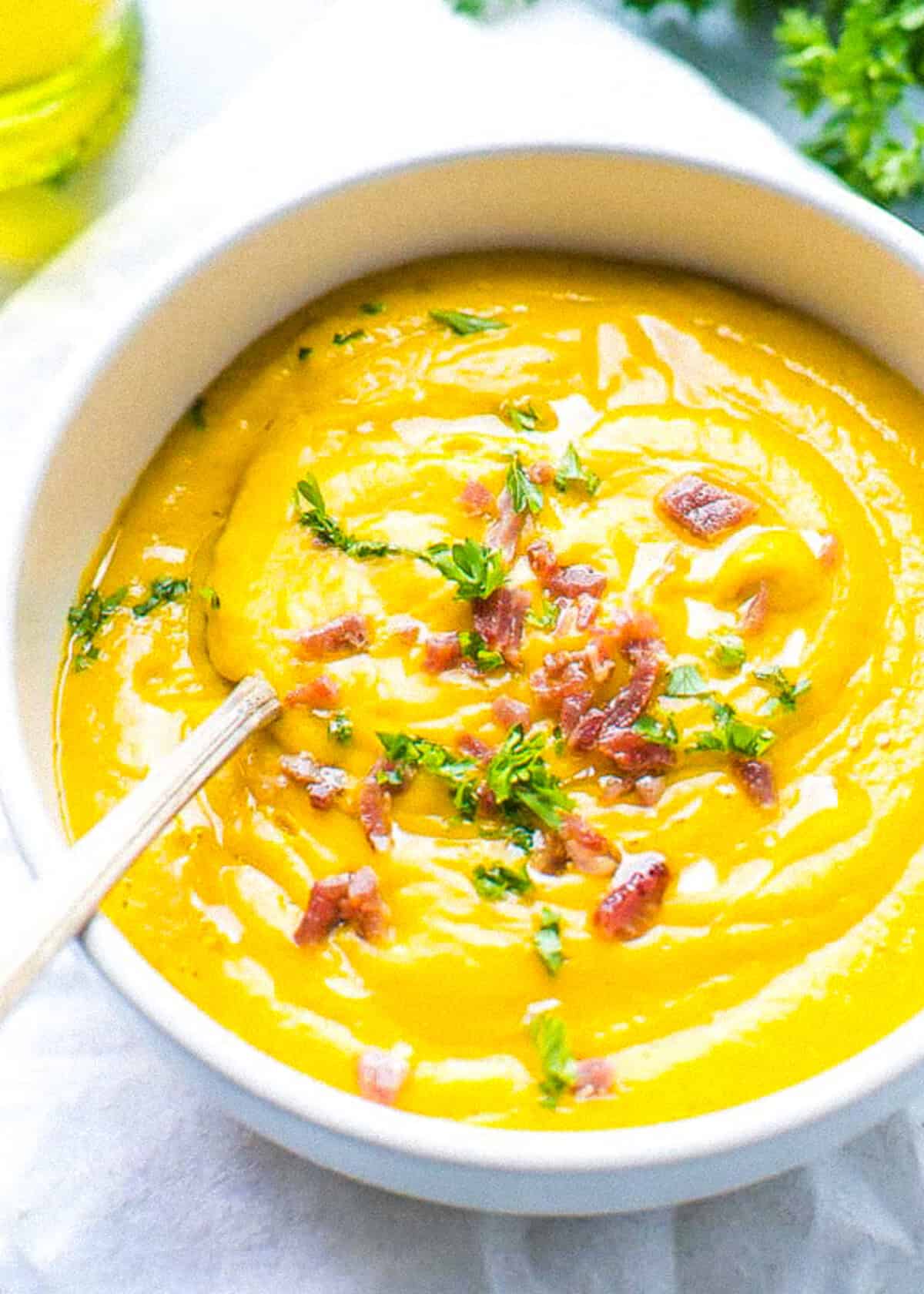 A white creamic bowl with creamy sweet potato soup, rich orange color and topped with bacon bits and chopped parsley. A bowl filled with creamy sweet potato soup. Made with sweet potatoes cooked in the microwave. Topped with bacon bits and parsley.