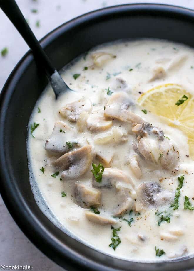 A bowl with white, lemony Creamy Mushroom Soup Recipe (Wolfgang Puck Copycat). Spoon scooping the mushrooms. Topped with parsley and lemon slice.