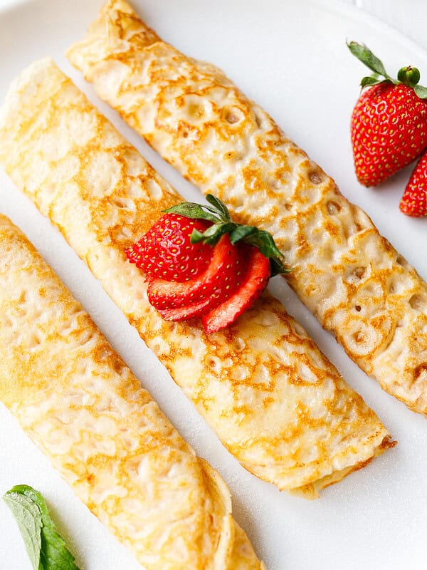 Three golden rolled crepes on a white plate topped with sliced fresh strawberries