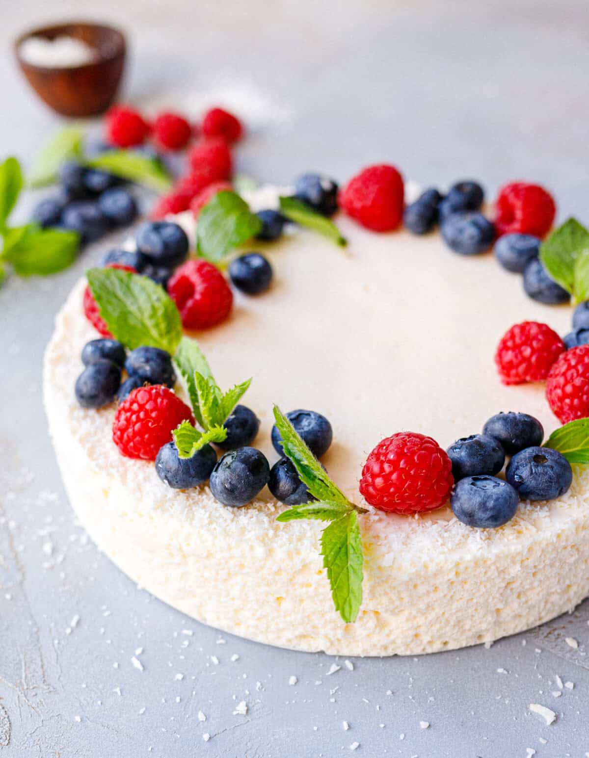 No bake yogurt cake topped with a wreath of raspberries, blueberries and mint, edges dusted with shredded coconut
