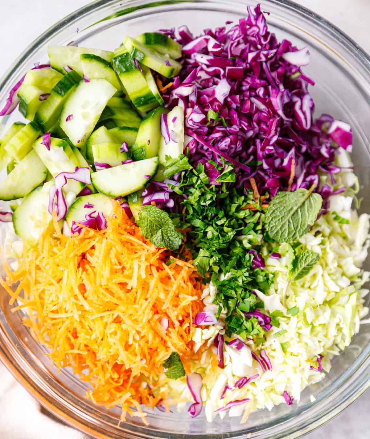 A clear glass bowl with purple, green shredded cabbage, carrots and cucumbers