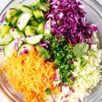 A clear glass bowl with purple, green shredded cabbage, carrots and cucumbers