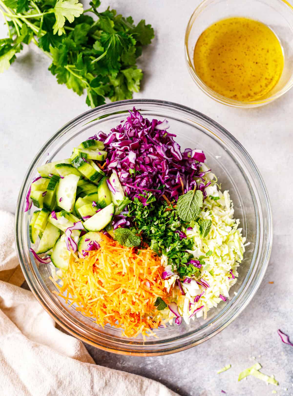 Chopped cabbage salad ingredients in a glass bowl with cucumber, carrots, purple cabbage, green cabbage, and fresh herbs