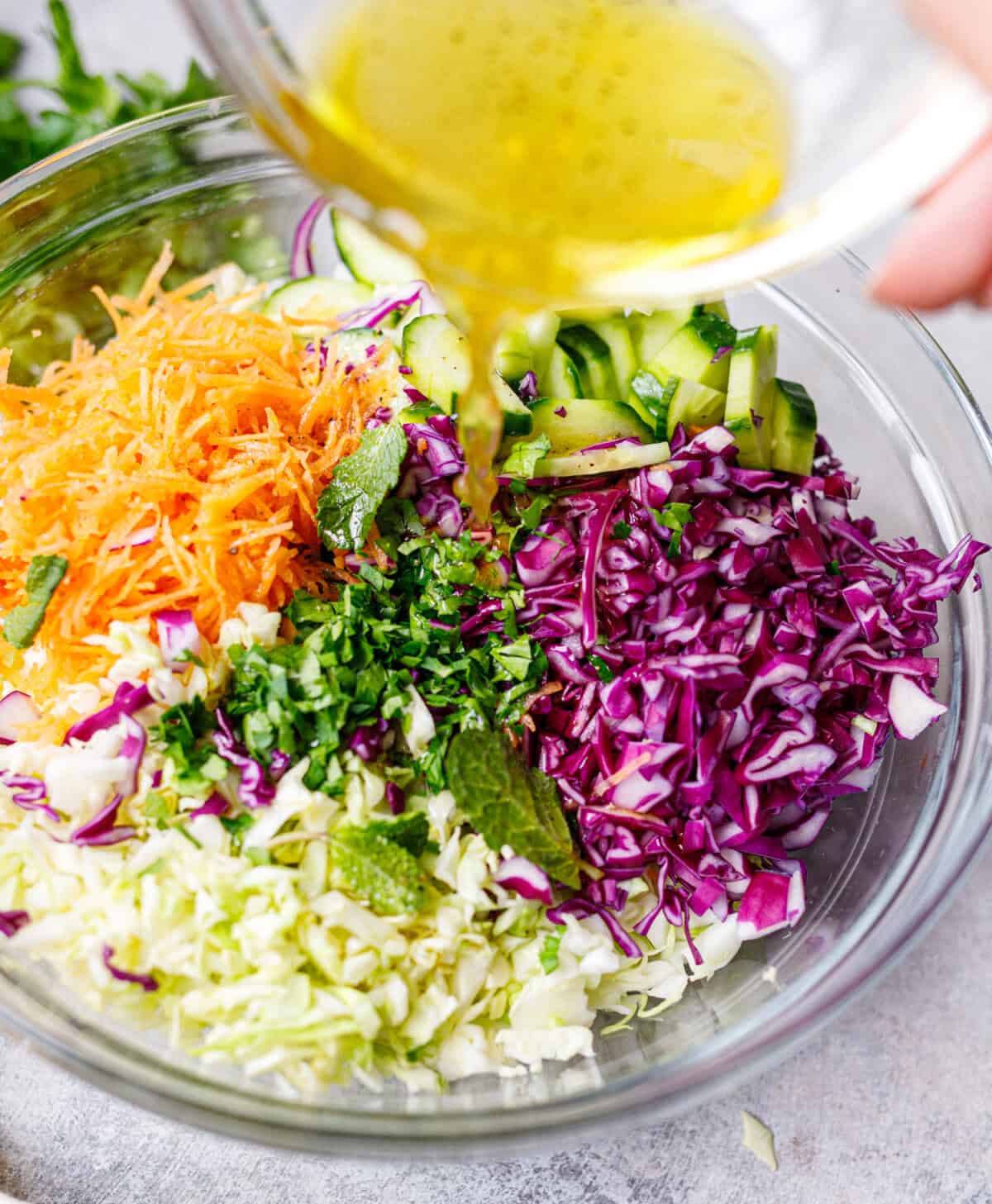 Pouring olive oil and vinegar dressing over cabbage salad ingredients in a glass bowl