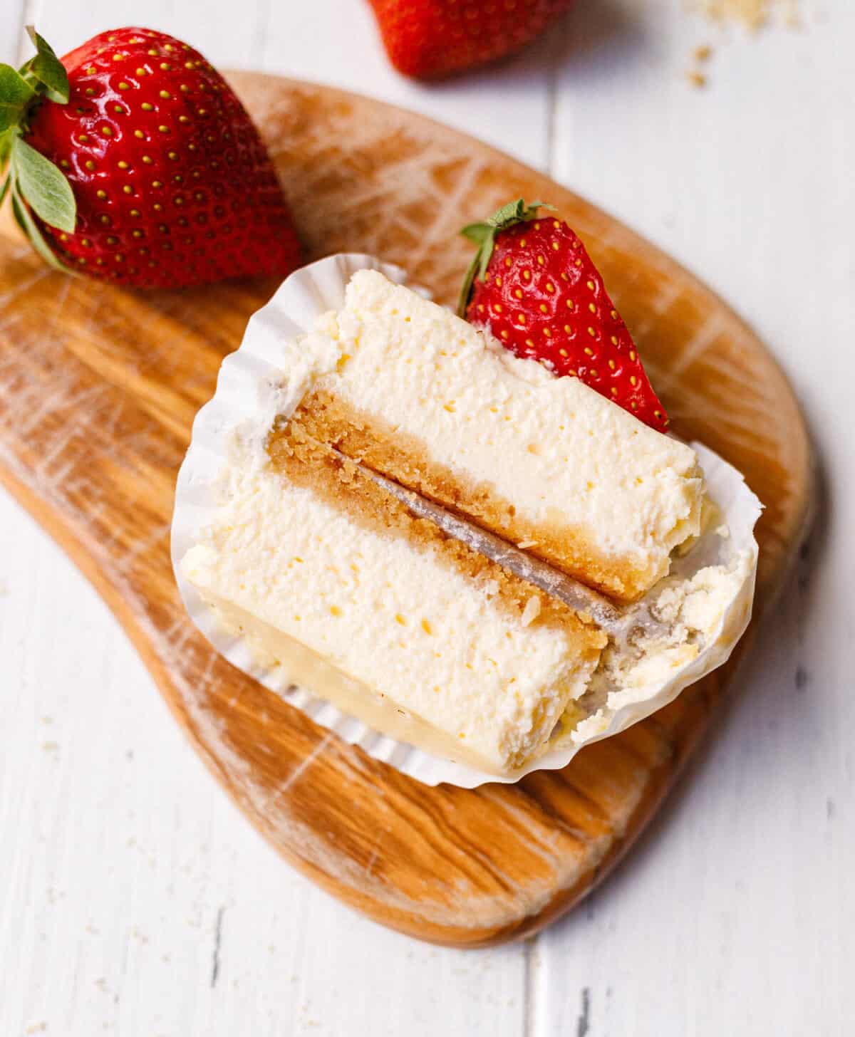 Cheesecake cupcake cut in half on a wooden board showing the creamy filling and graham cracker crust layer, with fresh strawberries nearby