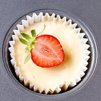 Mini cheesecake cupcake in a white liner in a dark muffin tin topped with a fresh strawberry half