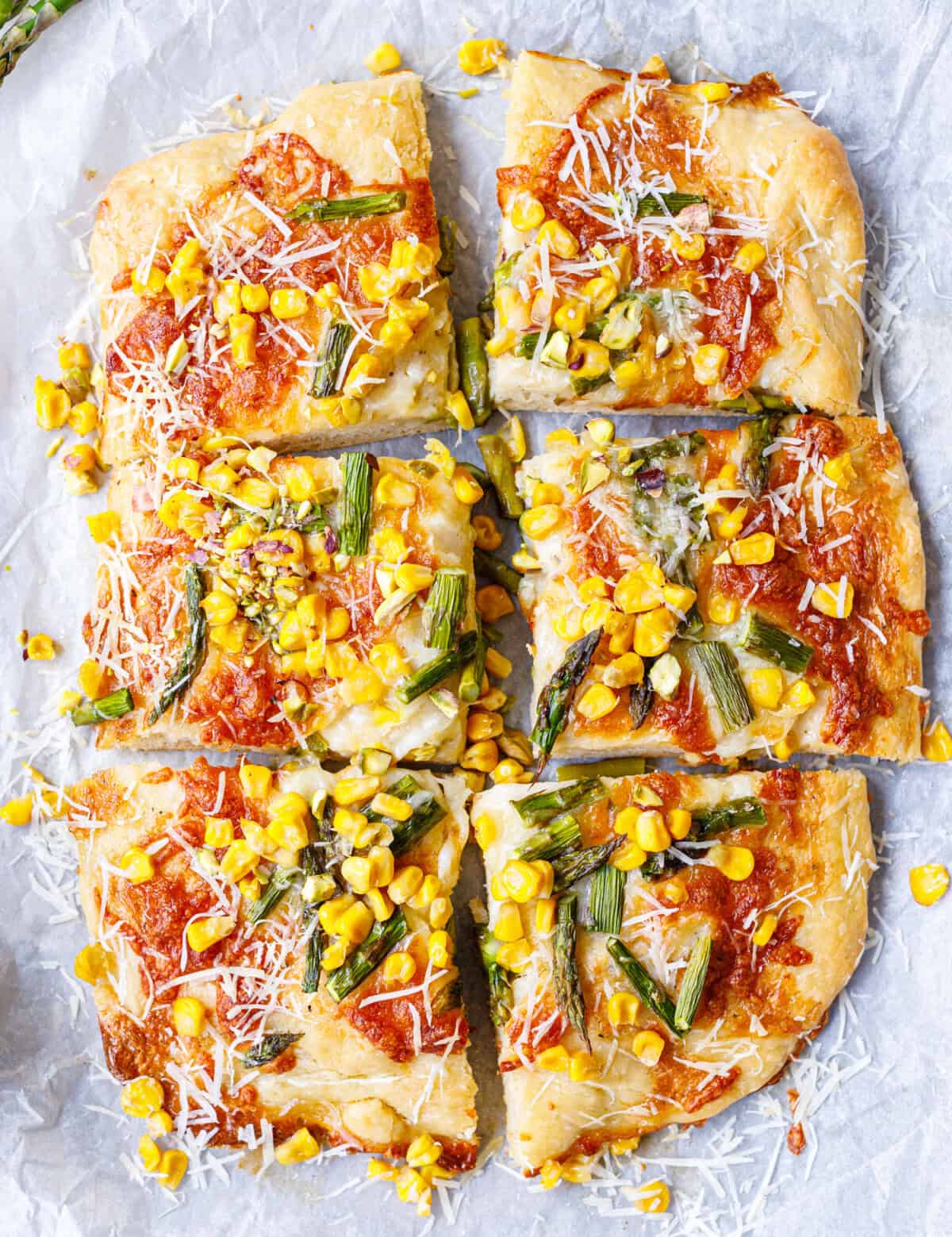 Whole asparagus corn pizza cut into 6 slices on parchment paper, topped with corn kernels, roasted asparagus, and shredded parmesan