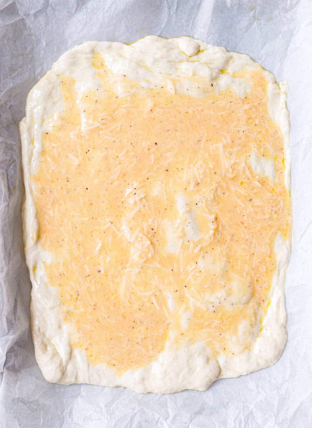 Pizza dough in pan with beaten egg and shredded mozzarella spread over the surface before baking