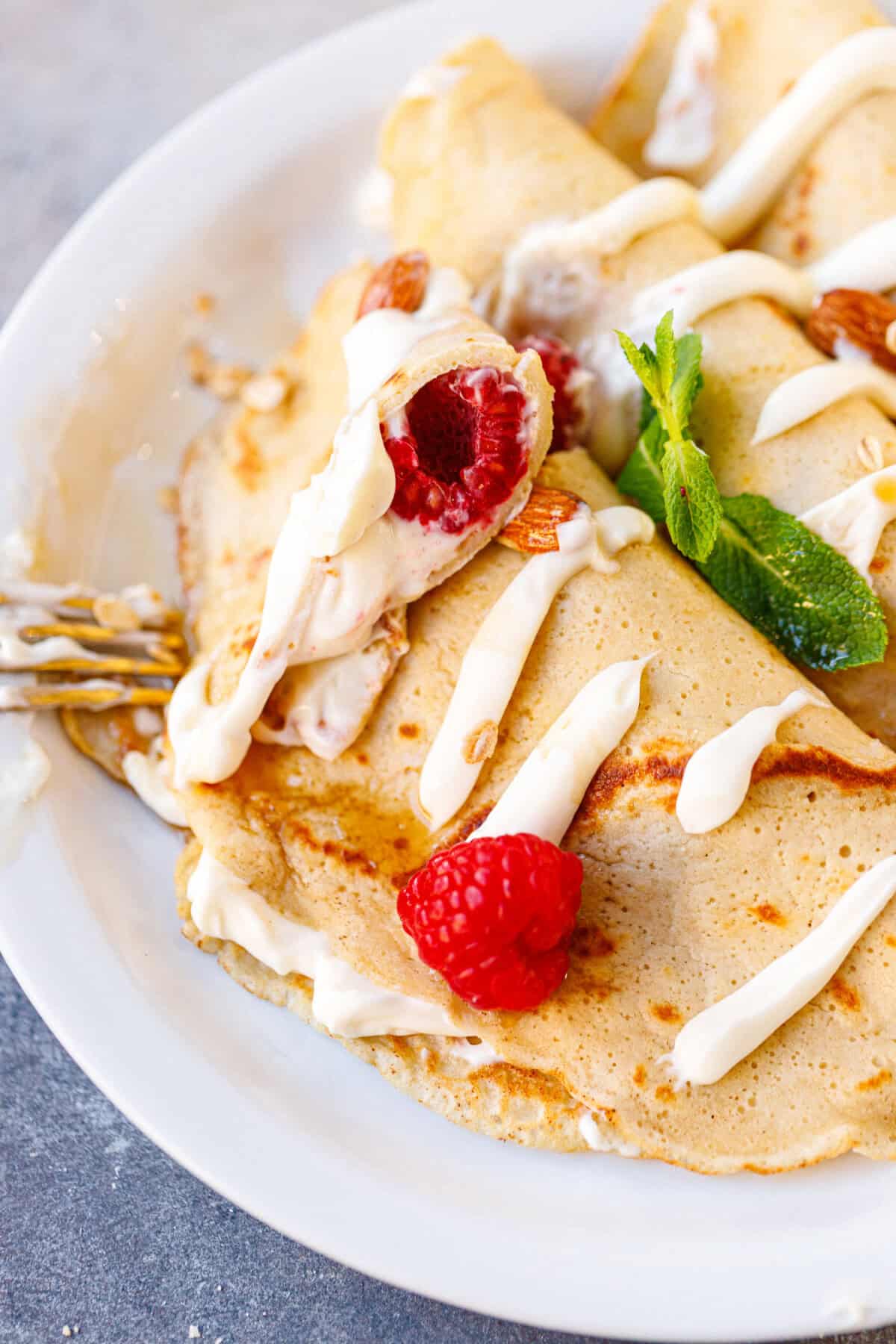 Folded almond oat cheesecake crepes on a white plate topped with raspberries, almonds, and cream drizzle