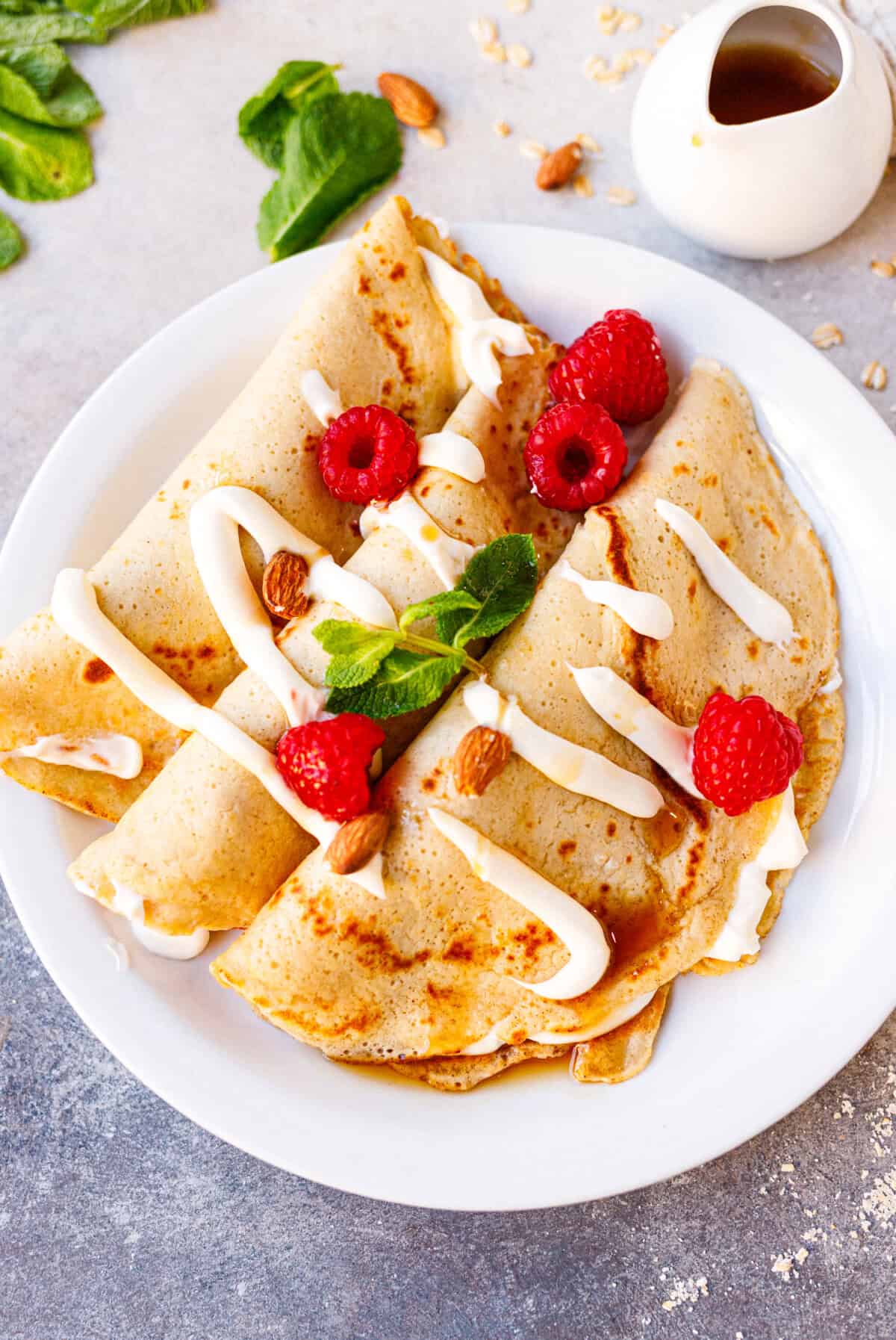 Four folded almond oat cheesecake crepes on a white plate topped with raspberries, almonds, and cream drizzle, with a small syrup pitcher in the background