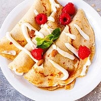 Four folded almond oat cheesecake crepes on a white plate topped with raspberries, almonds, and cream drizzle, with a small syrup pitcher in the background