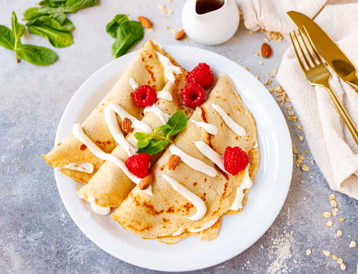 Almond oat crepes with cheesecake filling and raspberries on a white plate, styled with a gold fork and linen napkin