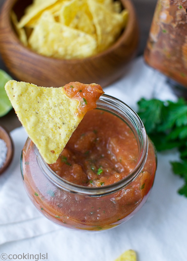 Easy blender salsa in a small glass jar with a bowl of tortilla chips on the side.