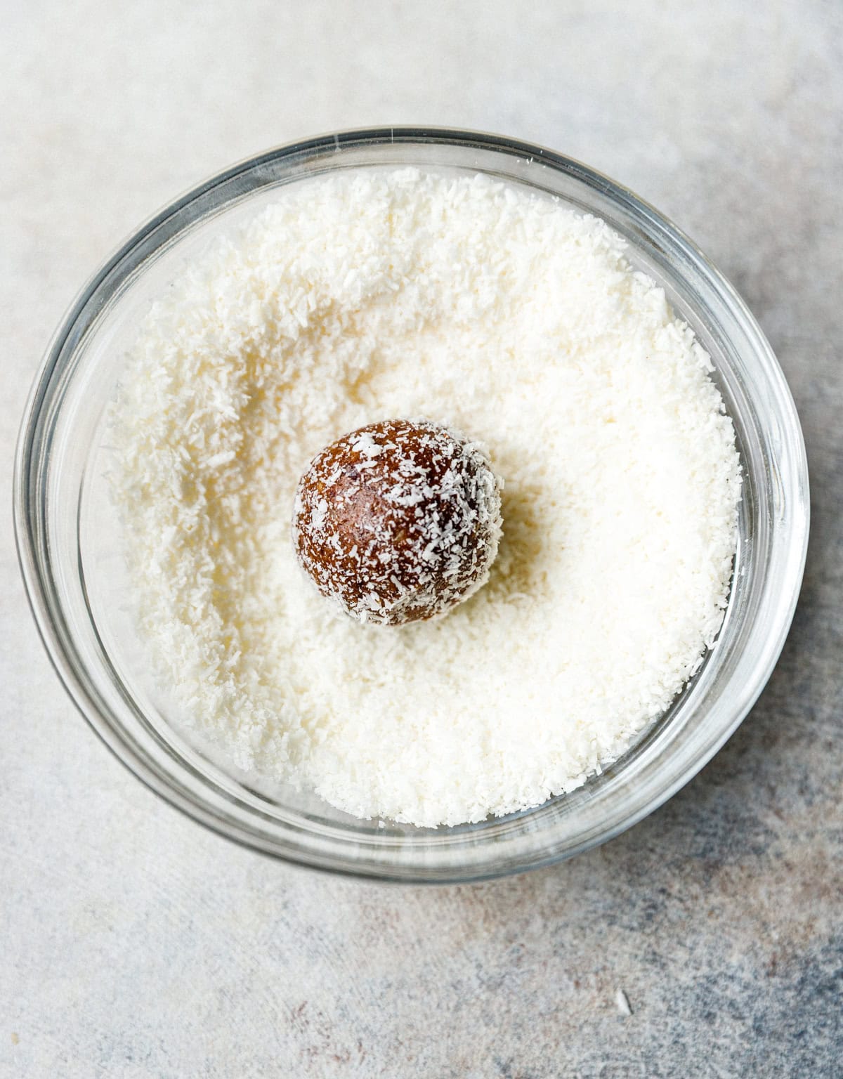 Homemade protein energy ball being rolled in shredded coconut in a glass bowl.