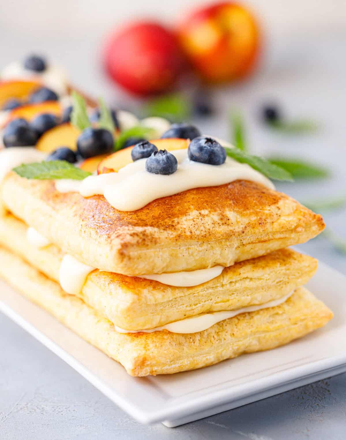 Overhead view of a finished puff pastry tart with vanilla cream, fresh nectarine slices, blueberries, and mint leaves on a white rectangular plate