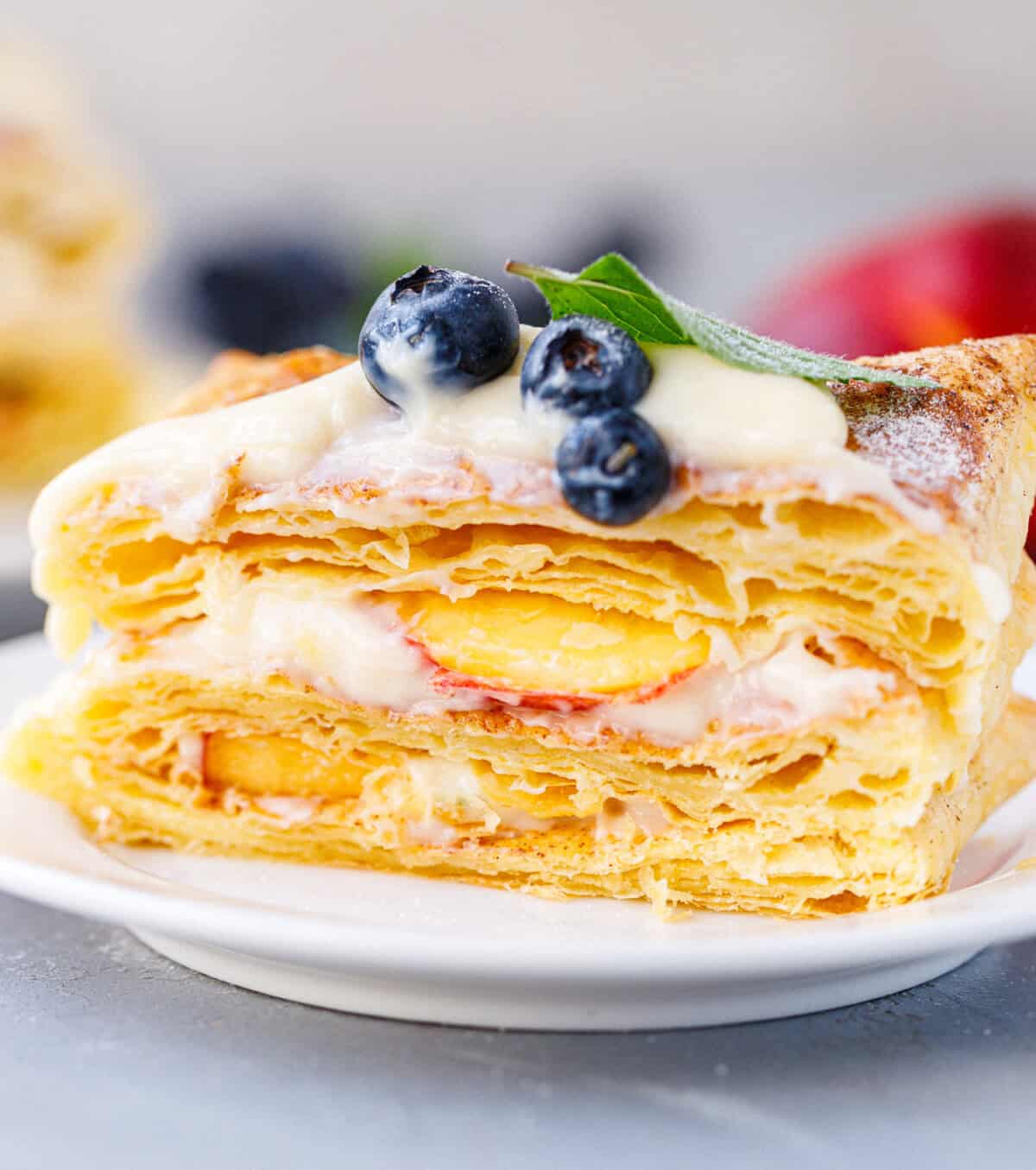 Single slice of puff pastry tart on a white plate showing flaky layers filled with vanilla cream and nectarine slices, topped with blueberries and mint