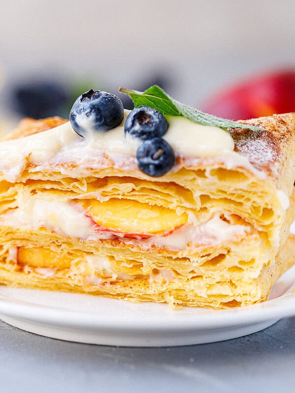 Single slice of puff pastry tart on a white plate showing flaky layers filled with vanilla cream and nectarine slices, topped with blueberries and mint