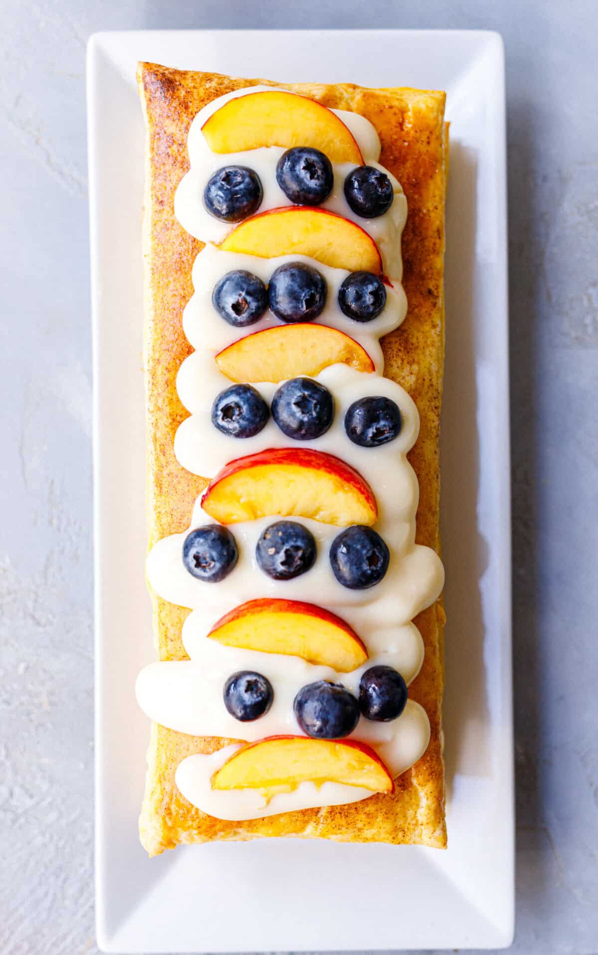 Overhead view of assembled puff pastry tart topped with vanilla cream, alternating nectarine slices and fresh blueberries on a white plate