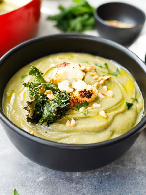 Roasted cauliflower kale soup in a black bowl, topped with roasted cauliflower, crispy kale, pine nuts and olive oil, with a red pot behind