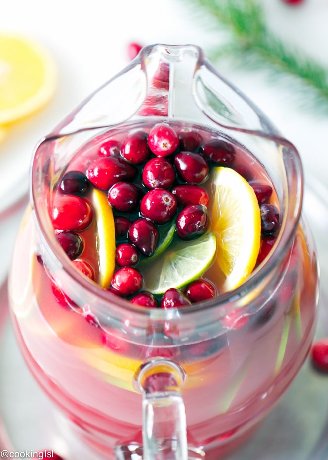 Cranberry Orange Punch in a clear festive pitcher, topped with fresh cranberries, lime and lemon slices. In a holiday setting, great for Christmas and NYE.