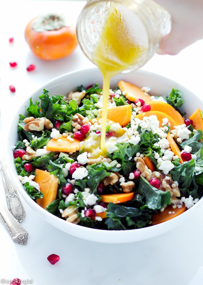 A bowl with kale persimmon salad, made with walnuts, feta, millet and pomegranate. Lime dressing poured on top from a jar.