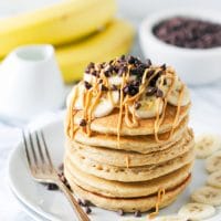 A stack of whole wheat peanut butter pancakes, on a plate, topped with sliced bananas, chocolate chips and peanut butter
